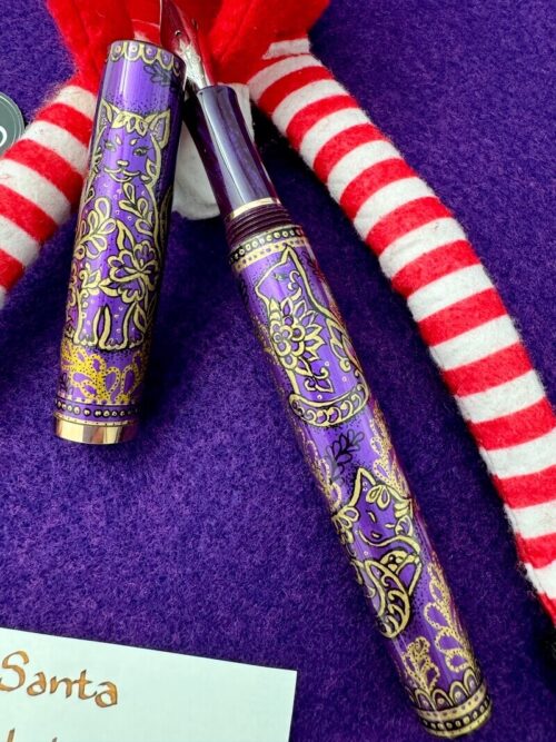 A fountain pen with drawings in gold of cats against a royal purple background. The pen is held by an Elf on the Shelf.